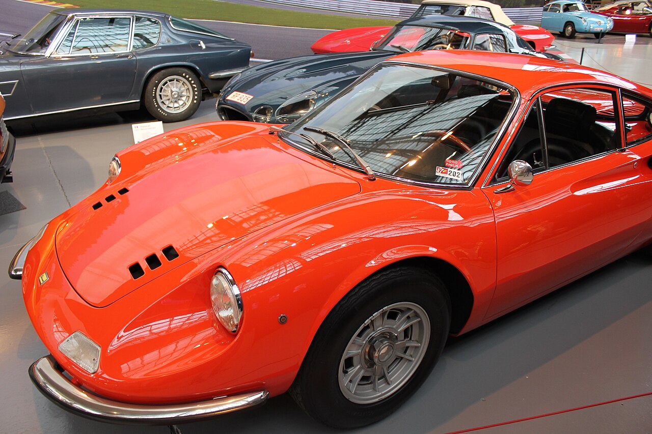 Orange-red Ferrari Dino 206 GT in a museum, front left three-quarter view from above, with other classic cars behind it.