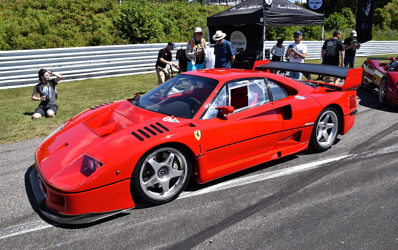 Red Ferrari F40 GTE competition car with a large rear wing, seen from the front left on tarmac with spectators nearby.