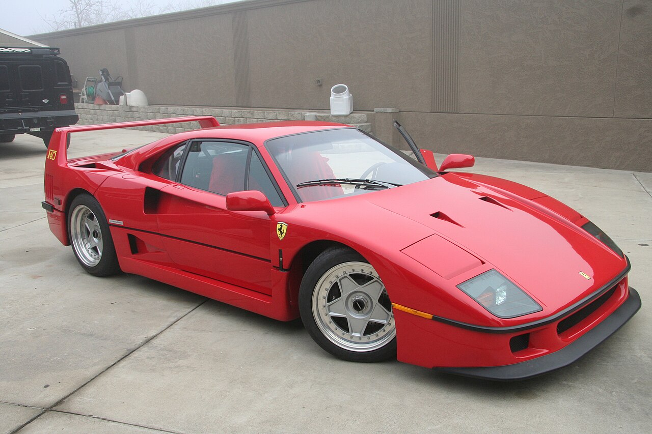 Red Ferrari F40 in front right three-quarter view, parked on concrete beside a wall on a misty day.