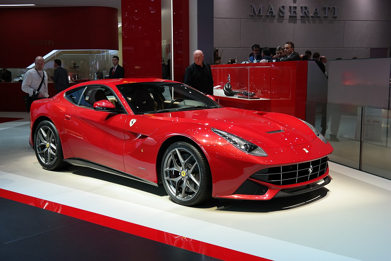 Red Ferrari F12berlinetta on a stand at the Geneva Motor Show, front right three-quarter view with visitors behind it.
