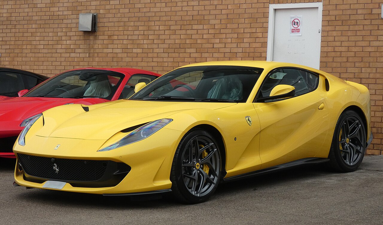 Yellow 2019 Ferrari 812 Superfast with dark alloy wheels, seen from the front left against a brick wall.