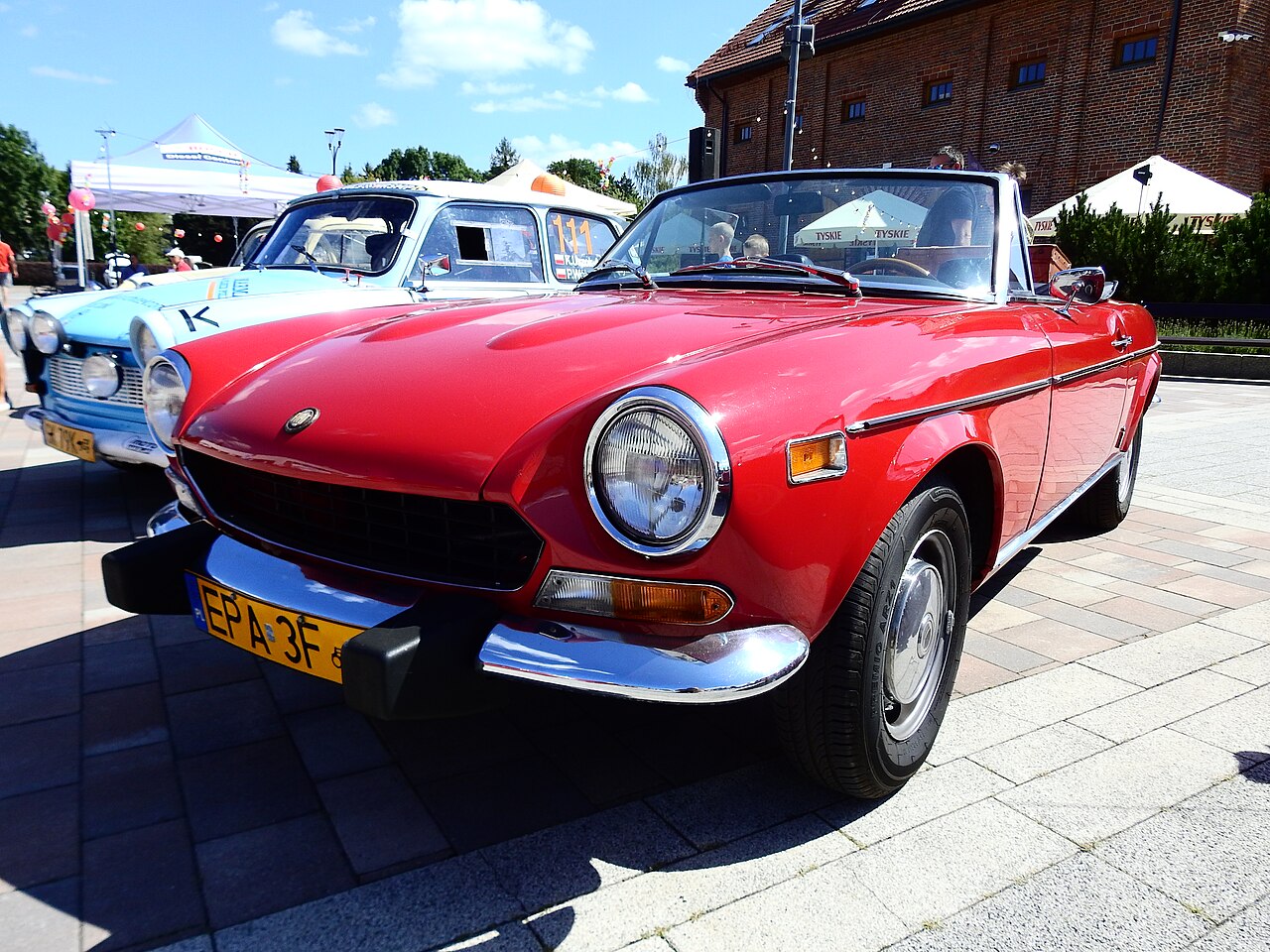 Red Fiat 124 Sport Spider with its roof down, low front left three-quarter view on paving at an outdoor classic car show.
