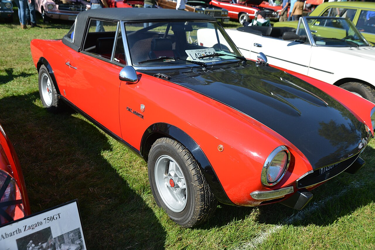 Red Fiat Abarth 124 Spider with a black bonnet and black roof, front right three-quarter view on grass at a classic car show.