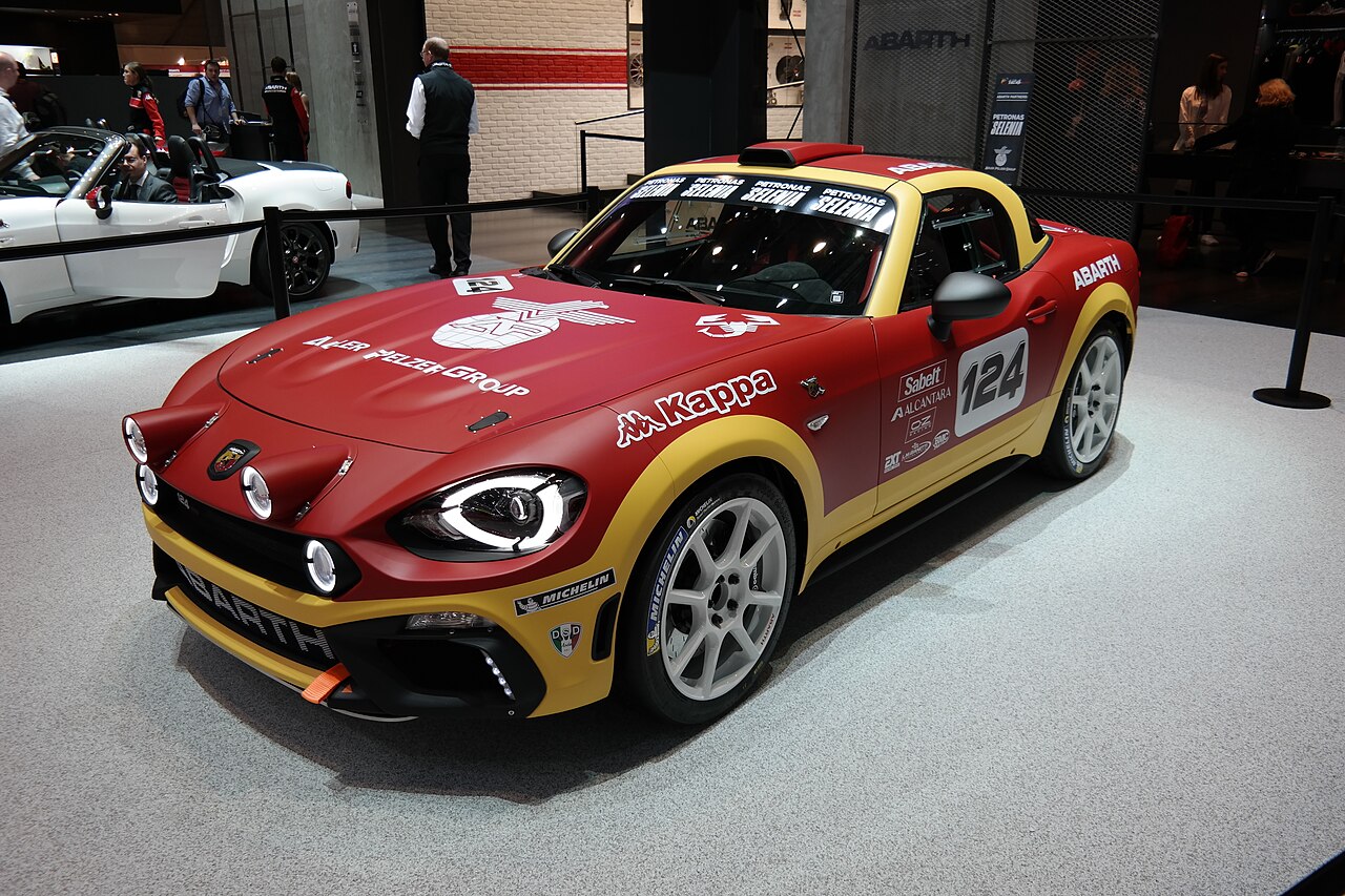 Red and yellow Abarth 124 rally car with sponsor decals, front left three-quarter view on a display stand at the 2016 Geneva Motor Show.