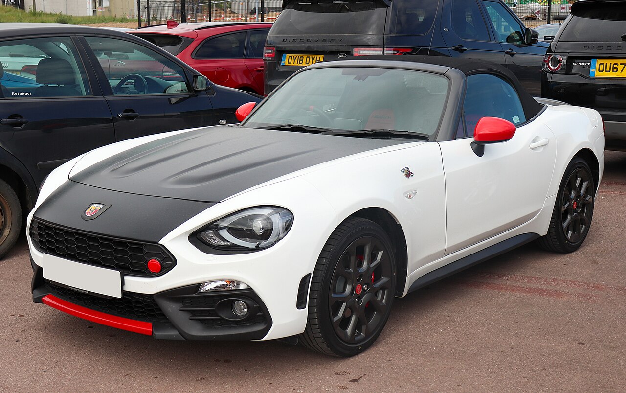 White 2018 Abarth 124 Spider with a matt black bonnet, red mirror caps and its soft top up, front left three-quarter view in a car park.