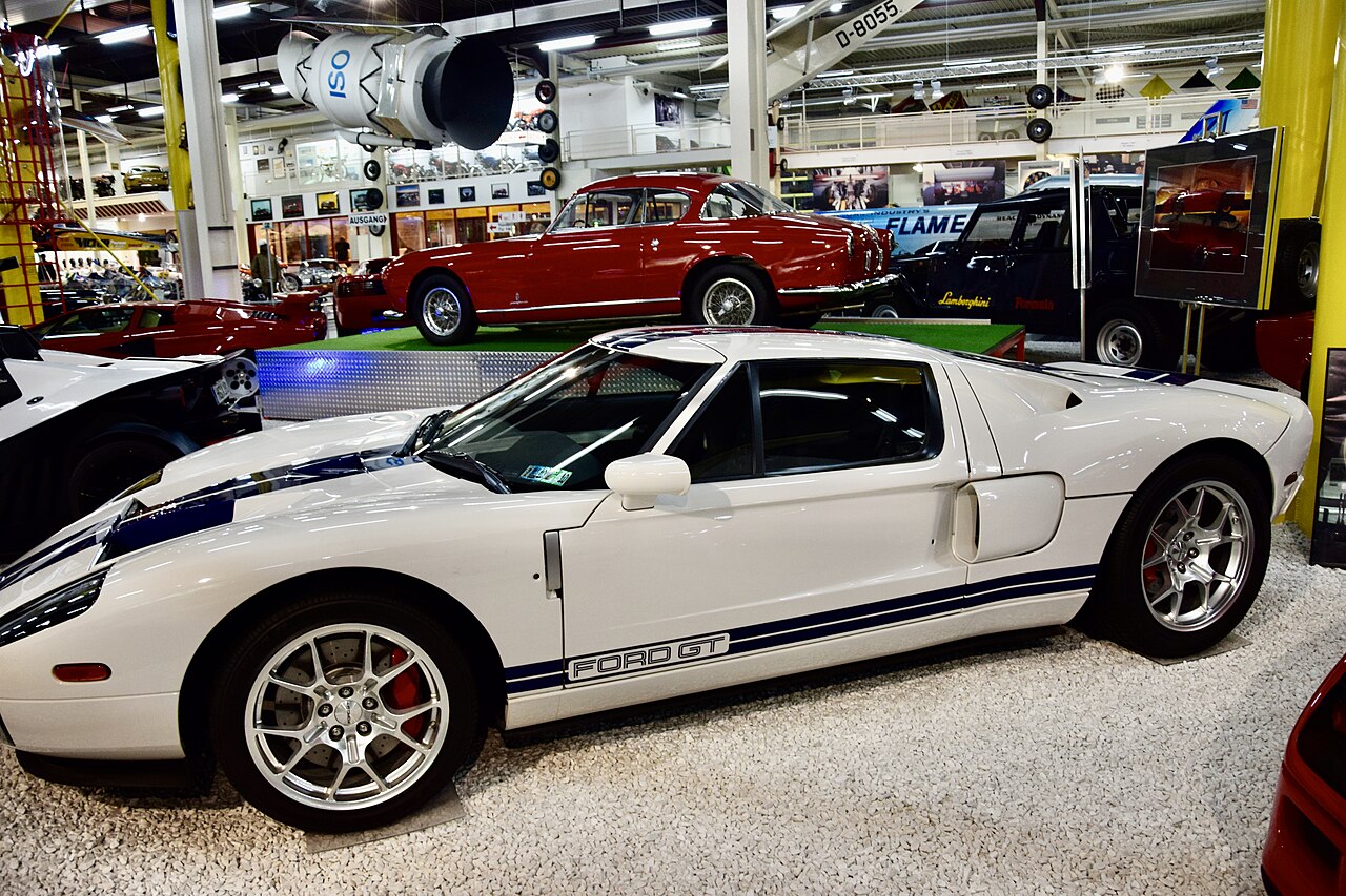 White 2005 Ford GT with blue stripes, side view in a museum with other cars behind it.