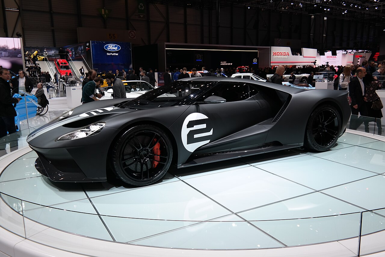 Matte black Ford GT with a white number 2 roundel on the door, side view on a glass platform at a motor show.