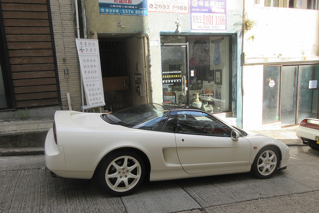 White first-generation Honda NSX with white alloy wheels, side-on with the nose to the right, parked on a narrow city street.