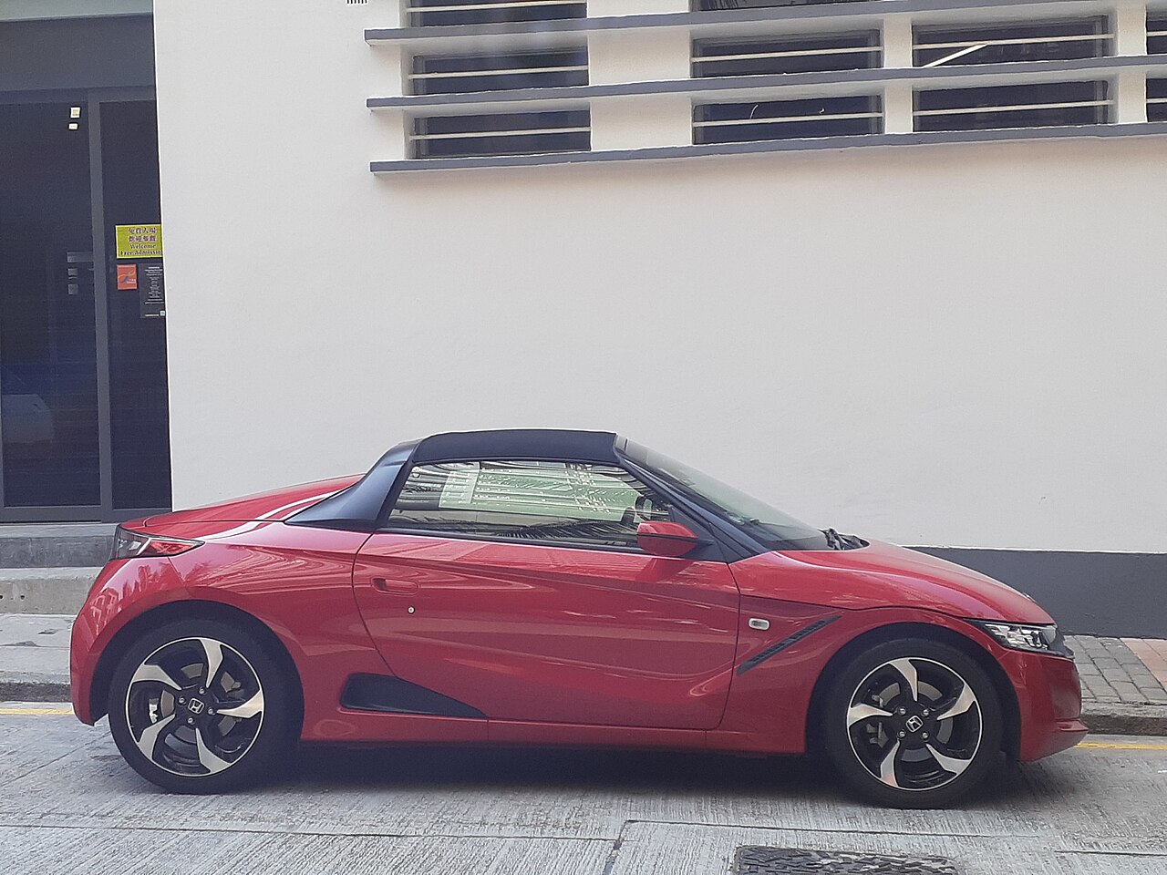 Red Honda S660 with its black soft top up, side-on view parked in front of a white wall.