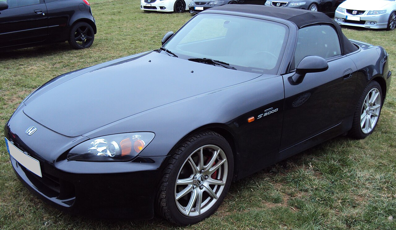 Black Honda S2000 with its soft top up, front three-quarter view parked on grass beside other cars.