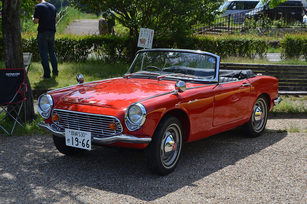 Red Honda S600 roadster with its soft top folded down, front three-quarter view parked on gravel.
