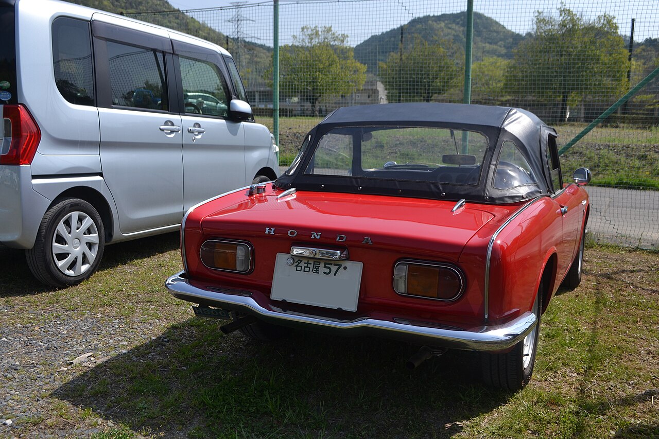 Red Honda S800 roadster with its soft top up, rear right three-quarter view on grass beside a silver minivan.