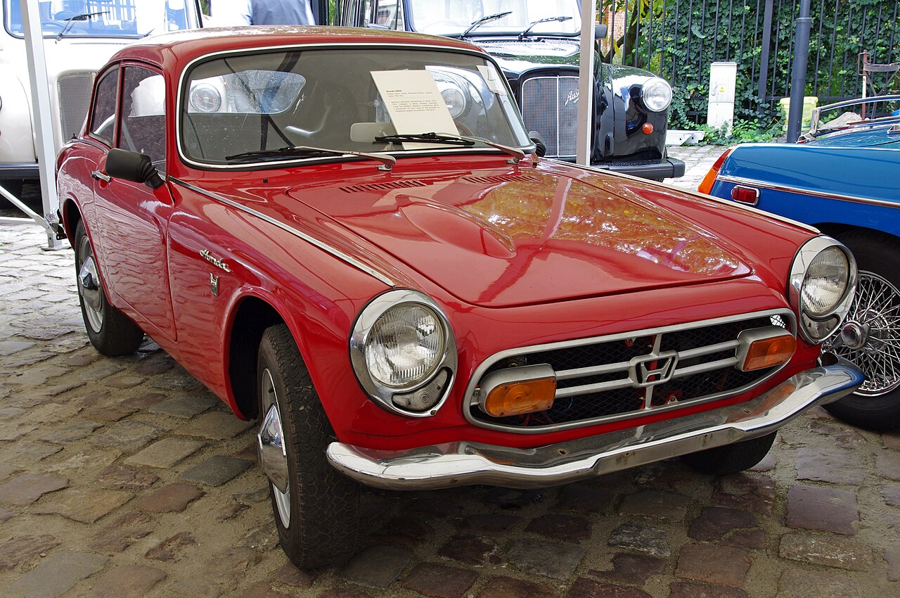 Red Honda S800 coupe, front three-quarter view on cobbles at a classic car show.
