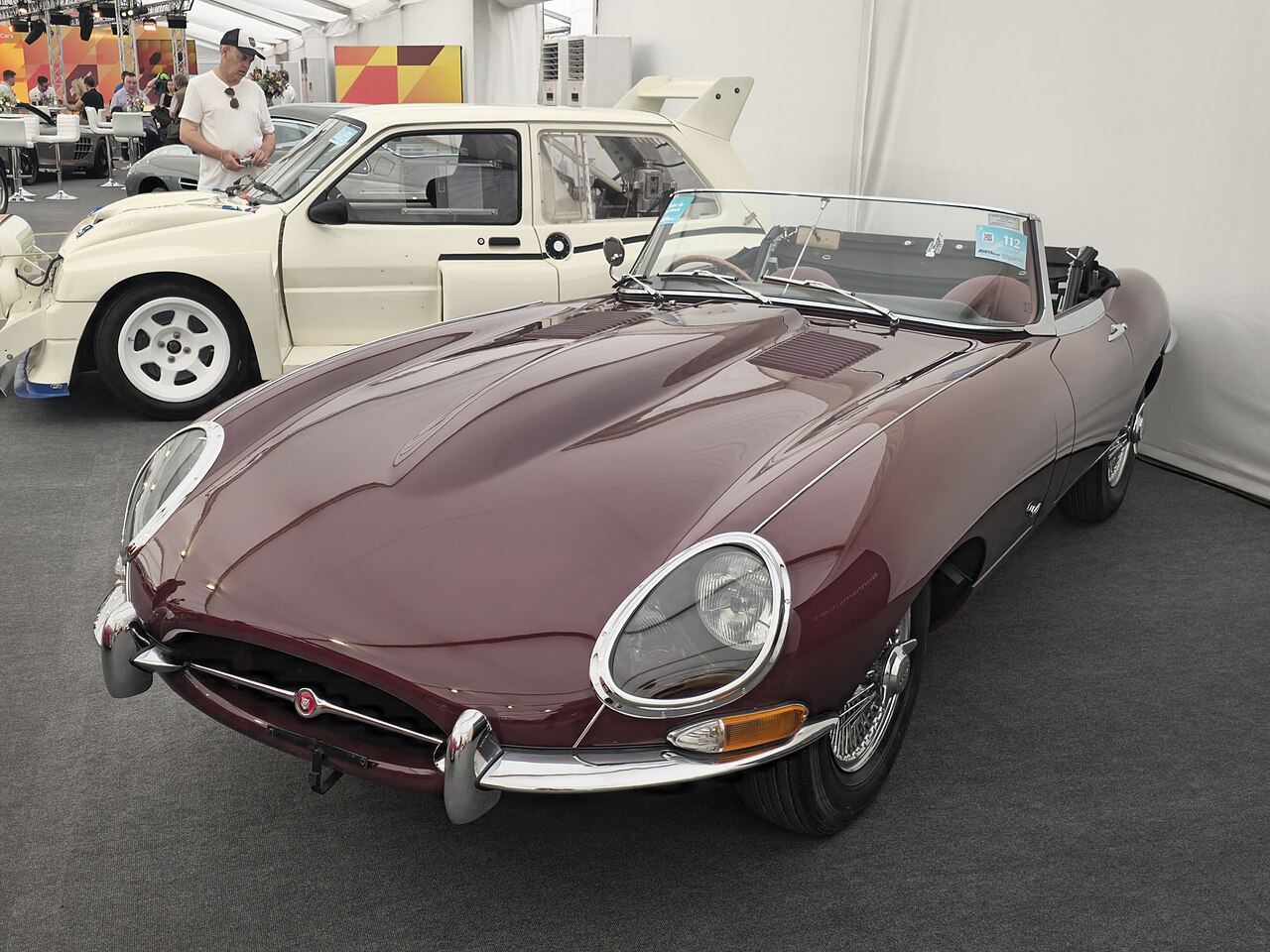 Maroon 1961 Jaguar E-Type roadster with its top down, front three-quarter view inside a show tent.