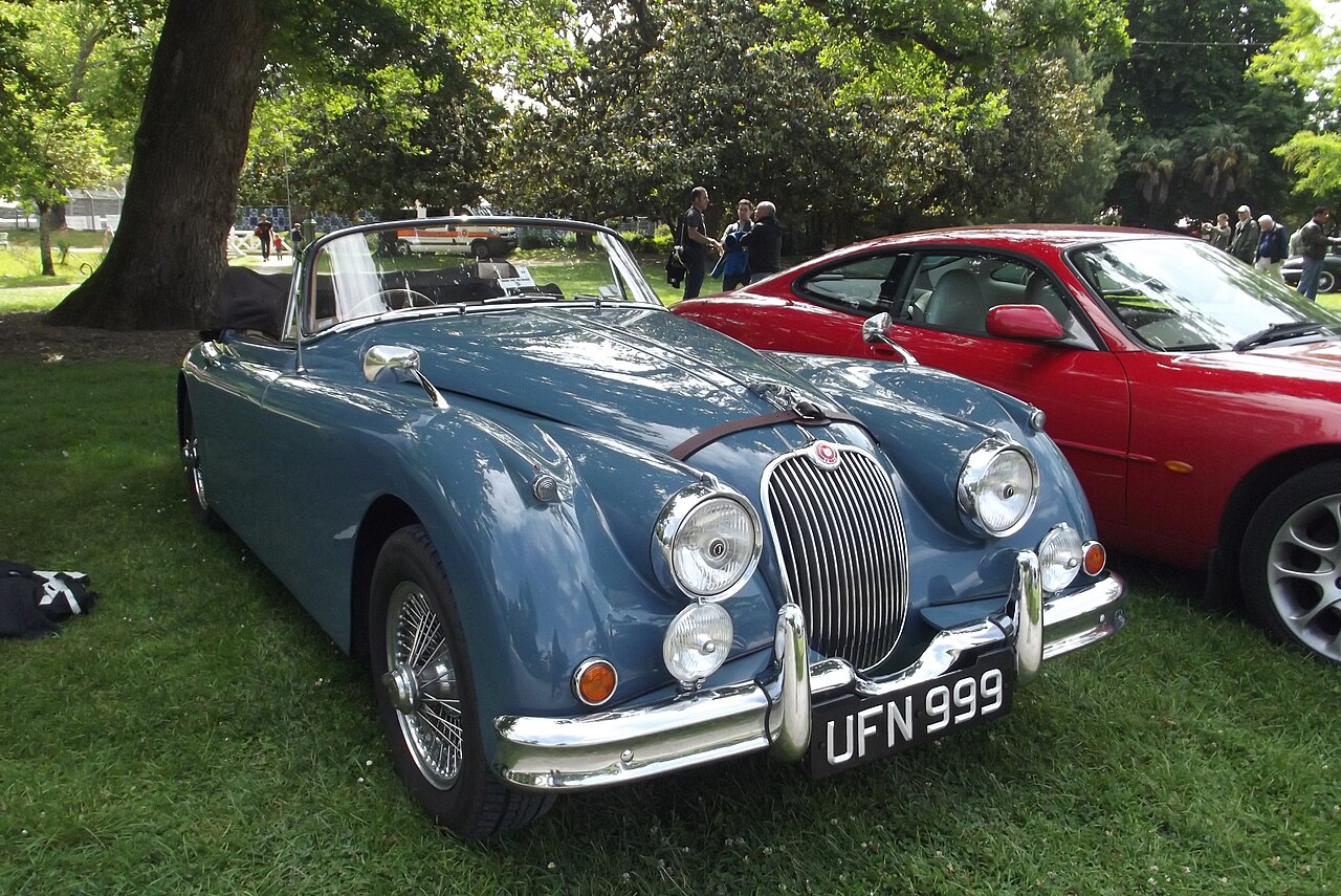Blue Jaguar XK150 drophead coupe with wire wheels and its hood folded down, parked on grass at a car show.