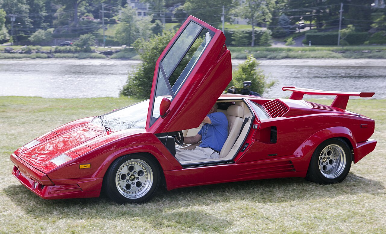 Red 1989 Lamborghini Countach 25th Anniversary with its scissor door raised, seen from the front left on a lawn by the water.