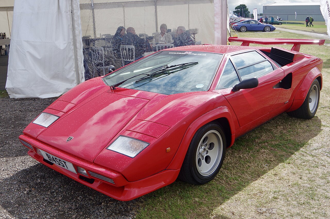 Red 1984 Lamborghini Countach with a rear wing, front three-quarter view with raindrops on the paint beside a marquee.