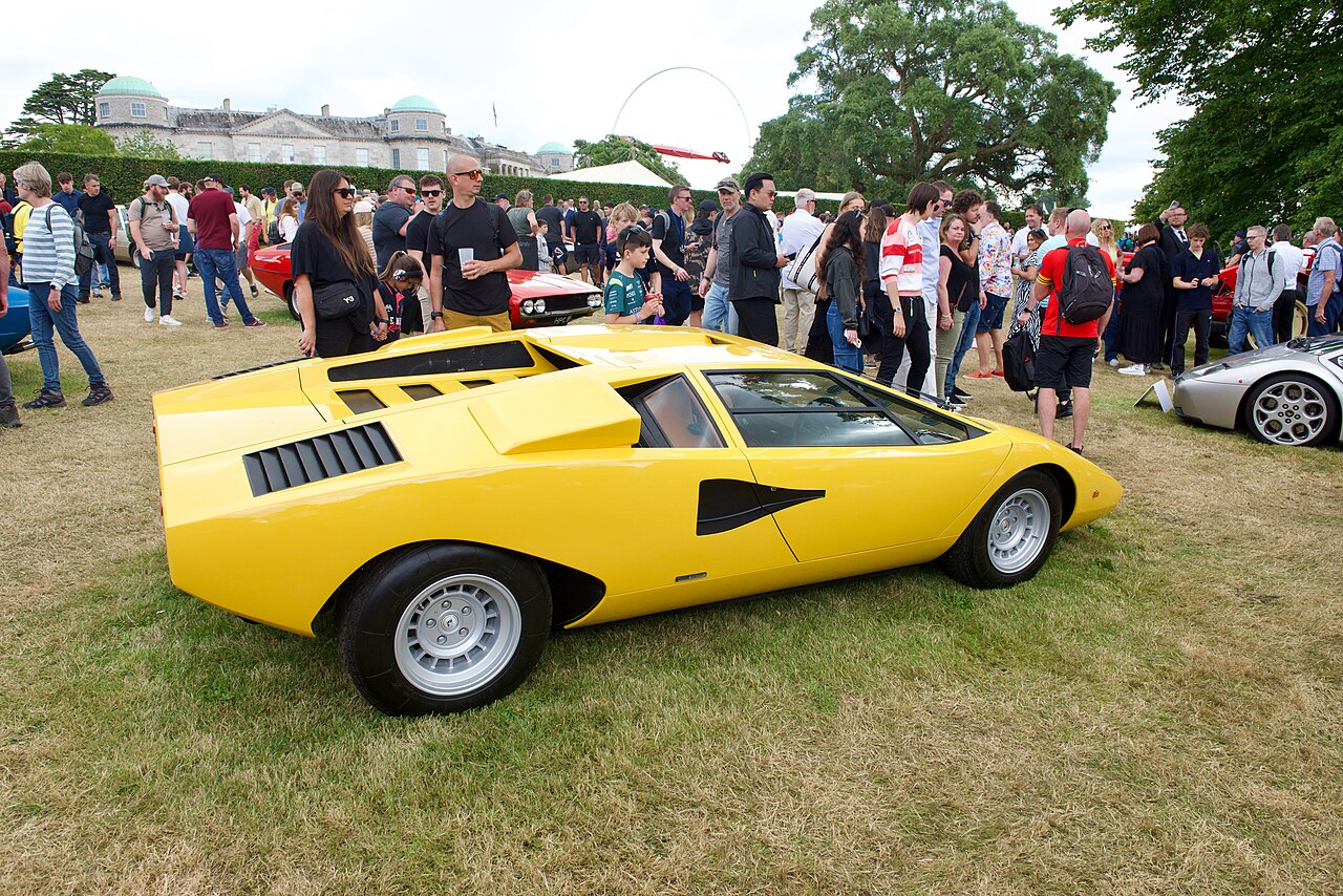 Yellow Lamborghini Countach seen from the side and rear on grass, with festival visitors gathered behind it.
