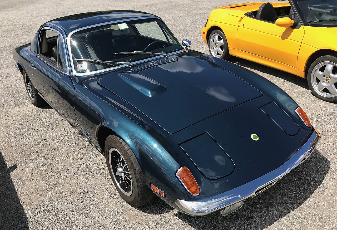 Dark teal Lotus Elan +2 coupe, front right three-quarter view from above on gravel, with a yellow Lotus beside it.