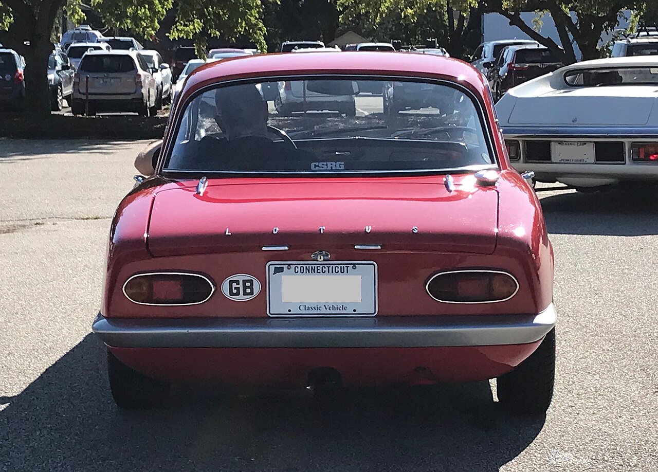 Red Lotus Elan S3 fixed-head coupe seen from directly behind in a car park, with a driver visible through the rear window.