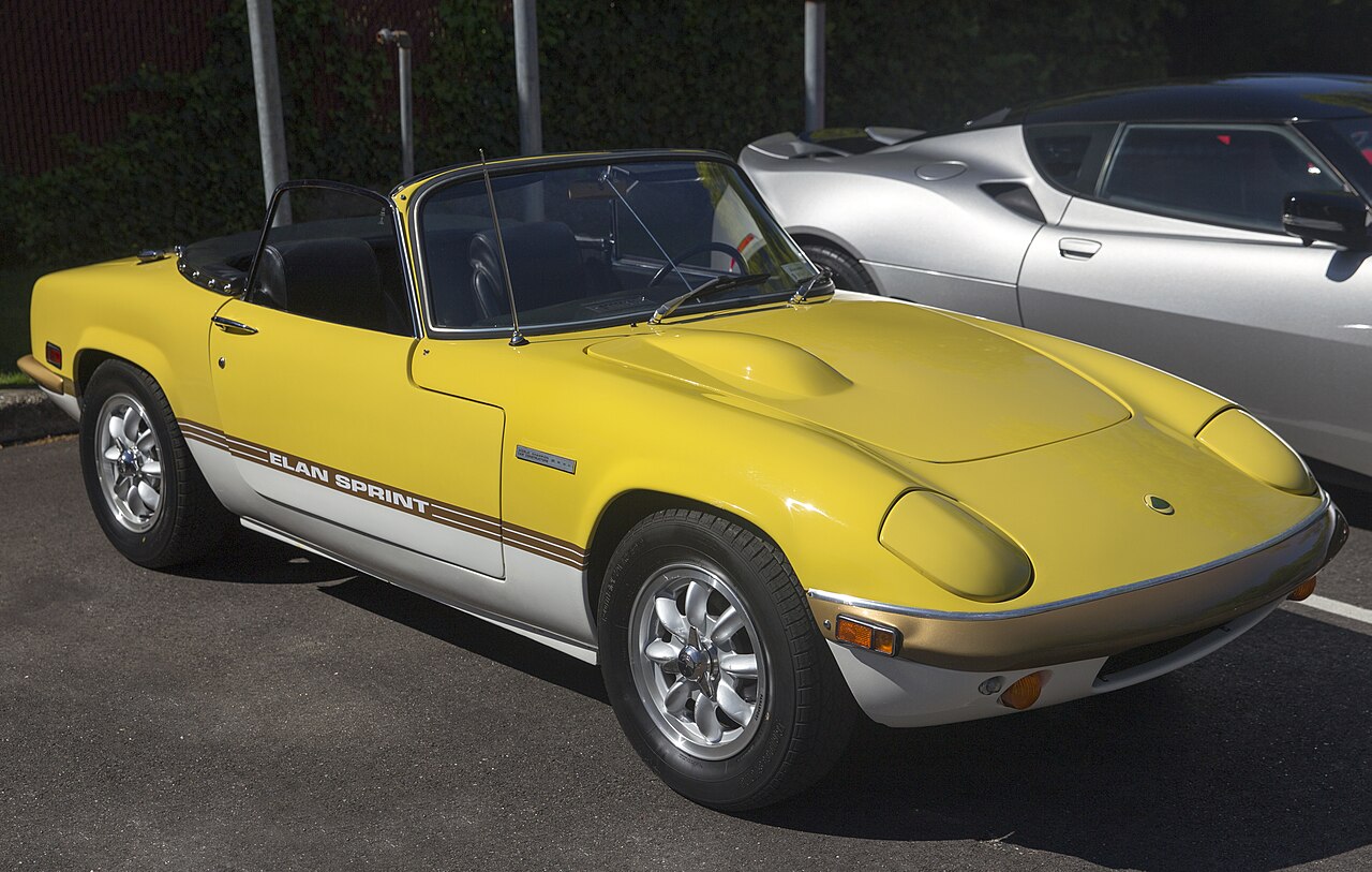 Yellow and white 1972 Lotus Elan Sprint roadster with its roof down, seen from the front right in a car park.