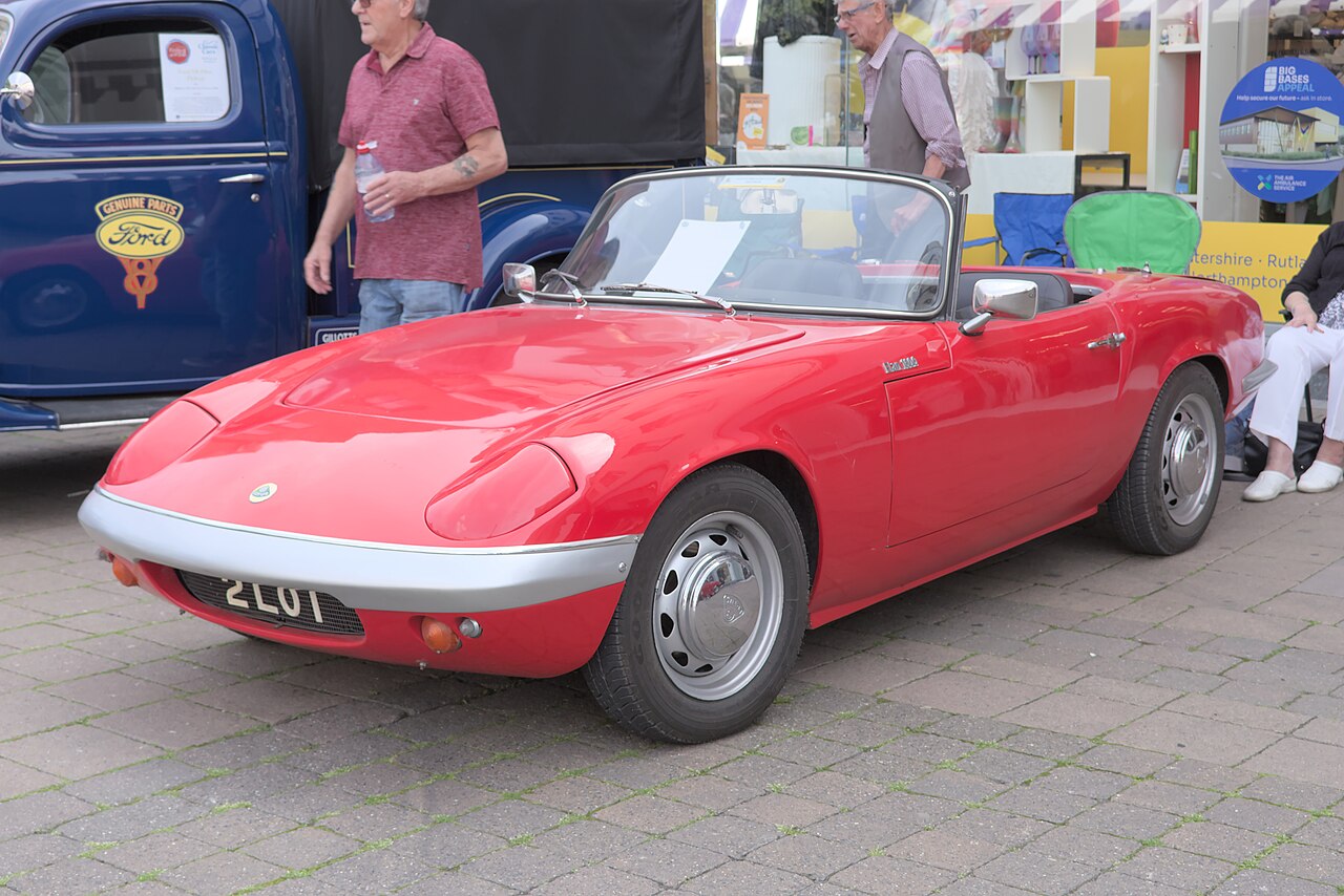 Red Lotus Elan roadster with its roof down, front left three-quarter view on a paved street at a car show.