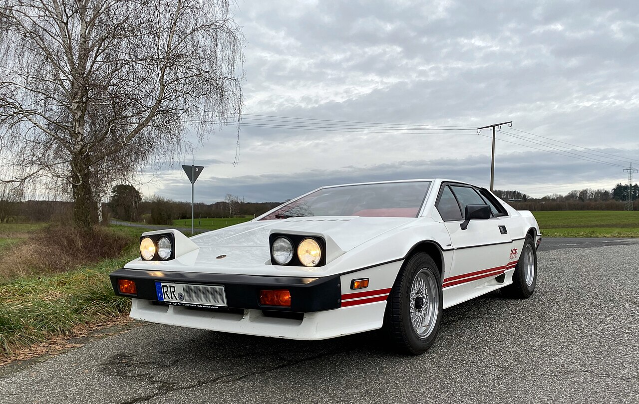 White Lotus Turbo Esprit with red side stripes and its headlamps raised, front left three-quarter view from a low angle on a country road.