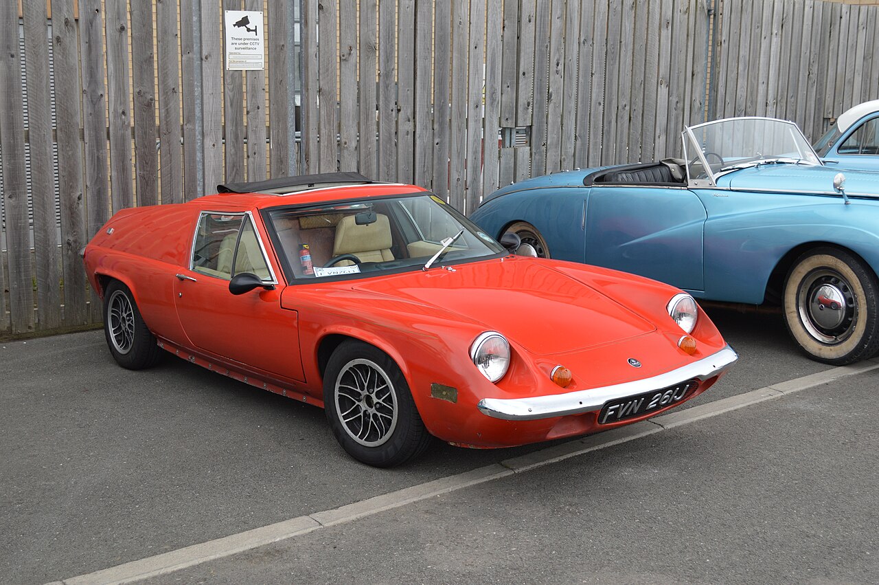 Orange-red Lotus Europa coupe, front three-quarter view in a car park beside a wooden fence.