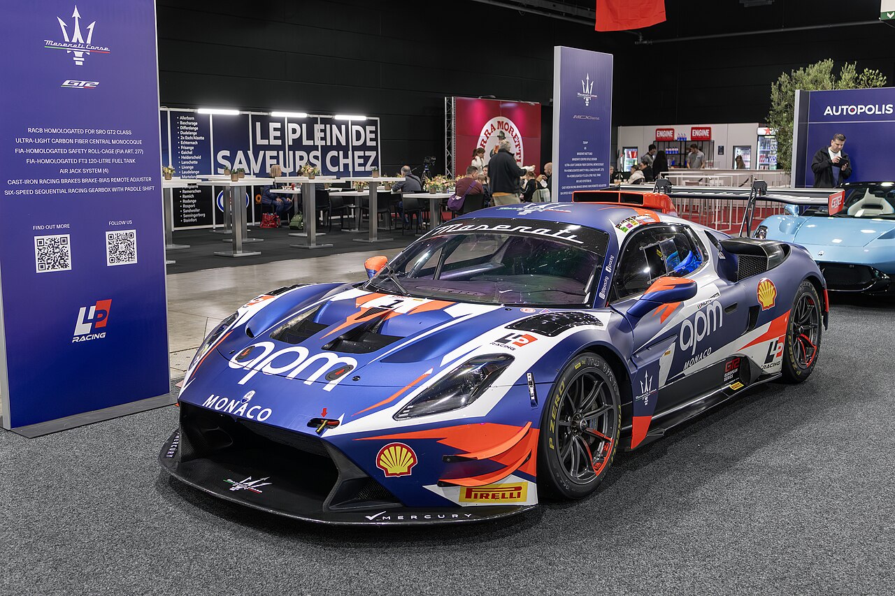 Maserati MC20 GT2 race car in blue, white and orange sponsor livery, front left three-quarter view on a motor show stand.