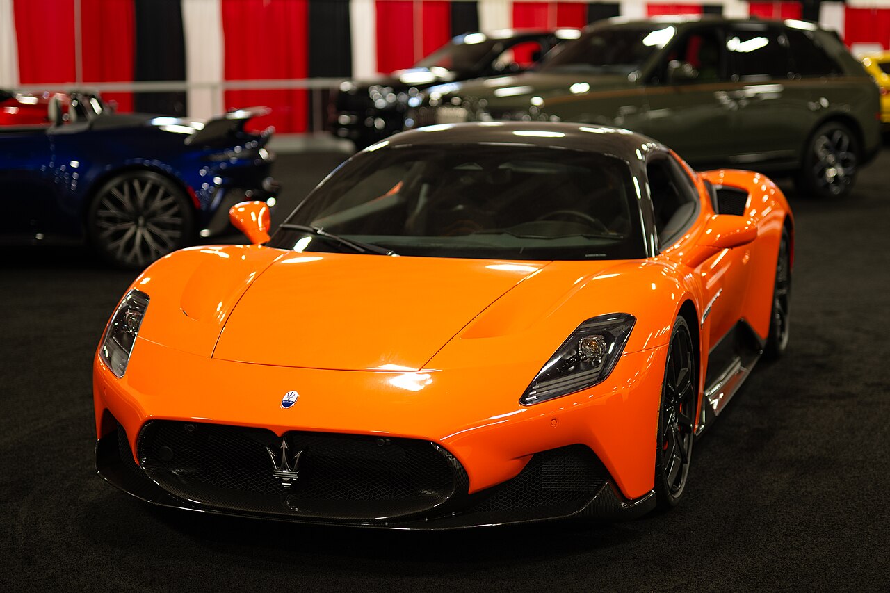 Orange Maserati MC20 with a black roof, front three-quarter view on a show-floor carpet.
