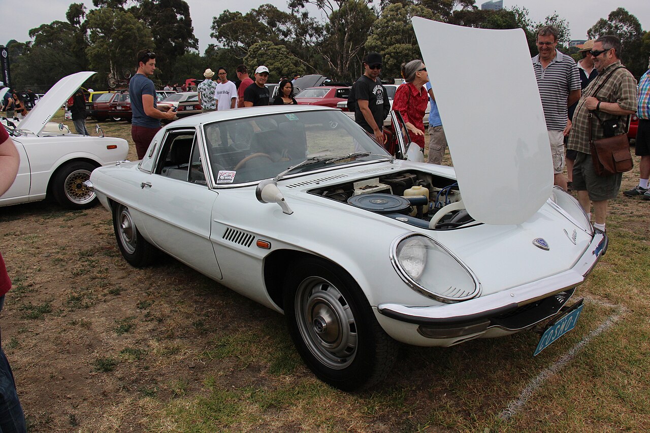 White 1967 Mazda Cosmo Sport with its bonnet raised, front three-quarter view on grass at a classic car show.
