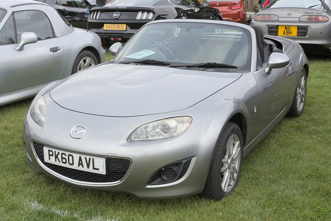 Silver Mazda MX-5 NC roadster with its roof down, front left three-quarter view on grass at a car show.