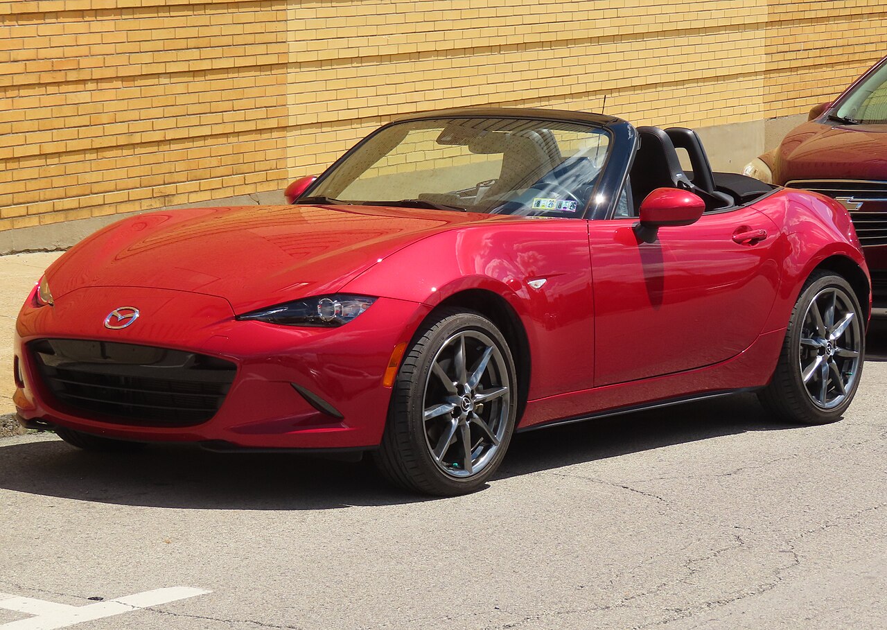 2016 Mazda MX-5 Miata Grand Touring in red with its roof down, front three-quarter view on a street beside a brick wall.