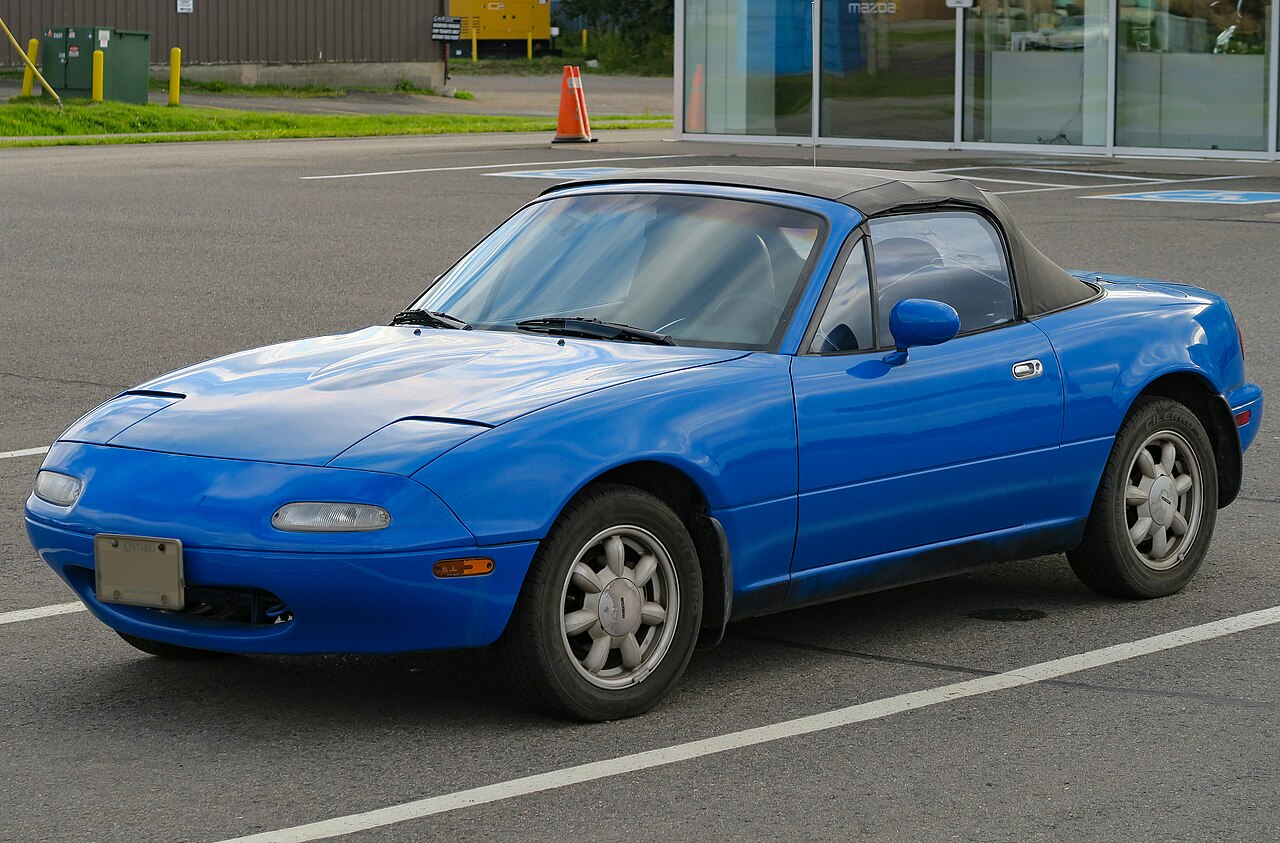 1990 Mazda MX-5 Miata in blue with its black soft top up, front three-quarter view in a car park.