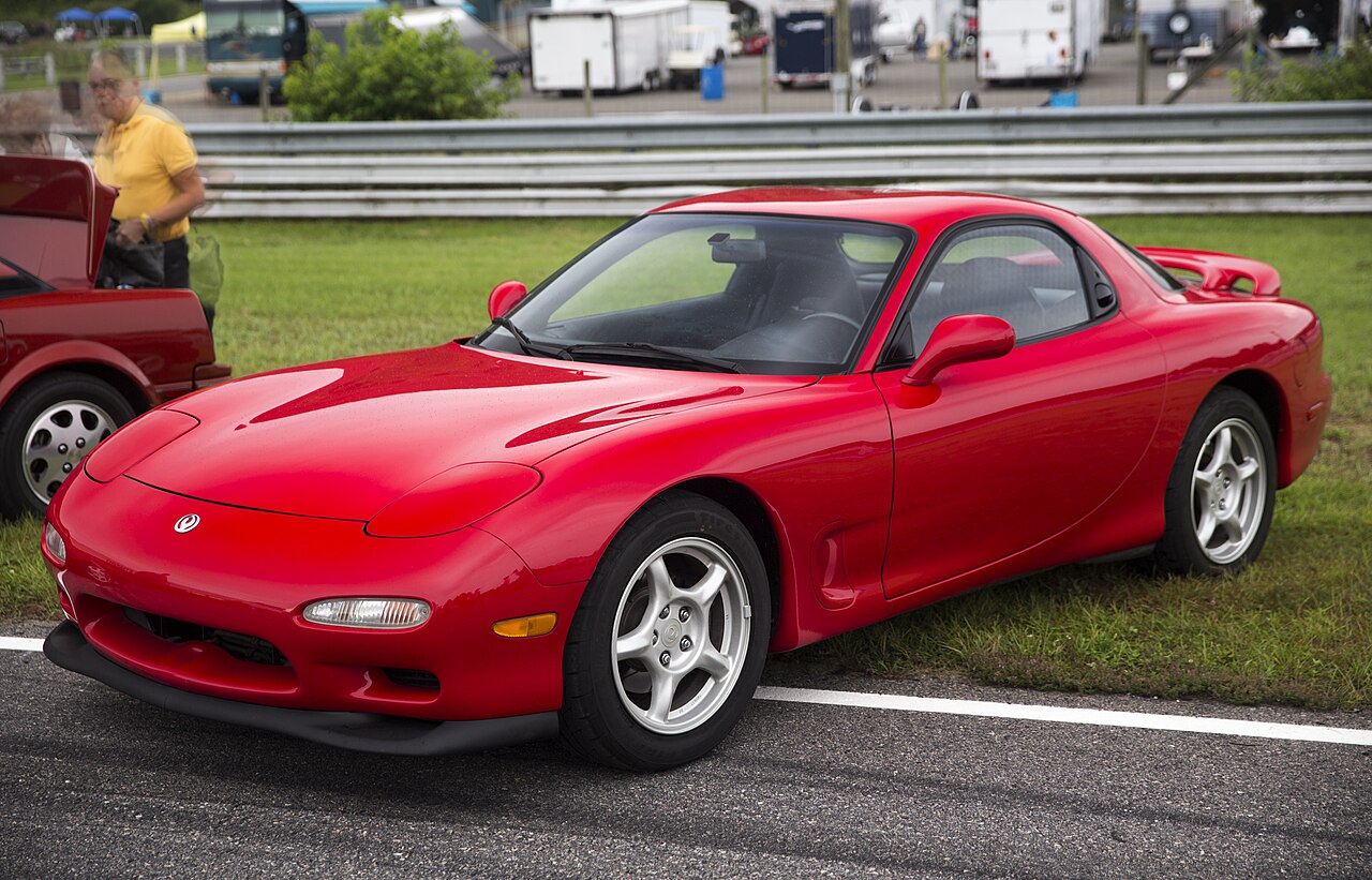 Red 1994 Mazda RX-7 R2 with silver wheels, seen from the front left on a grass verge at a race circuit.