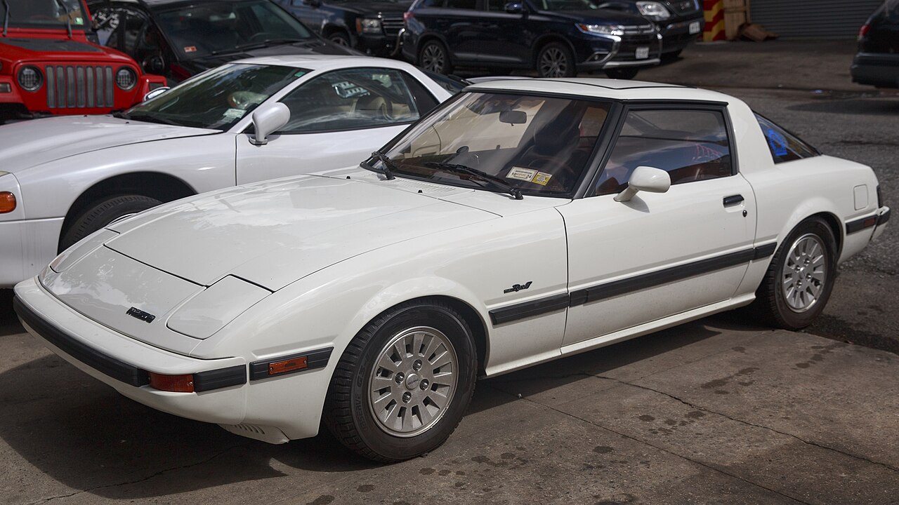 White 1985 Mazda RX-7 GSL-SE coupe, seen from the front left in a car park with other cars behind it.