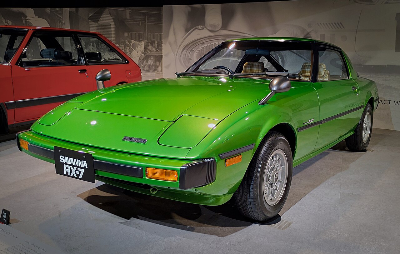 Green first-generation Mazda Savanna RX-7 coupe, seen from the front left on display in a museum.