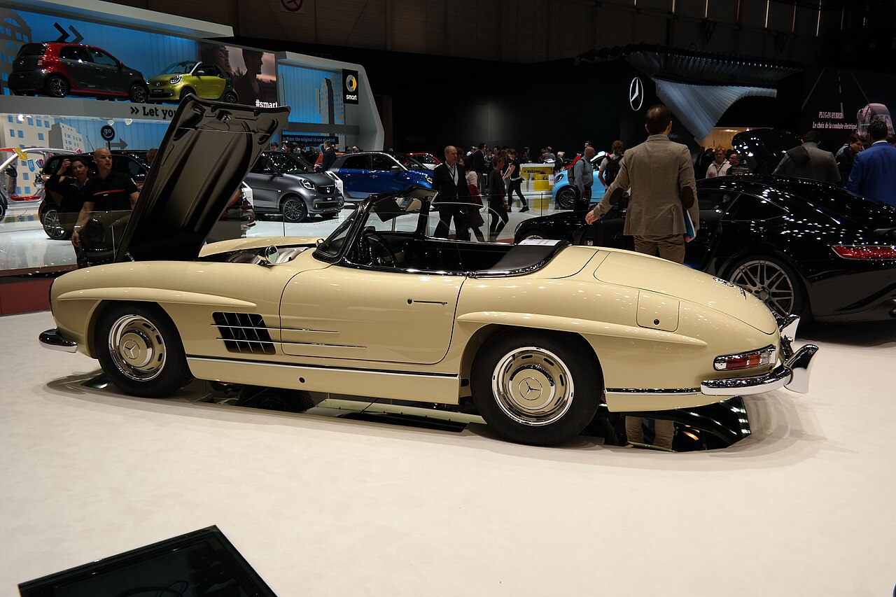 Beige Mercedes-Benz 300 SL Roadster with its roof down and bonnet raised, side-on view on a stand at a motor show.