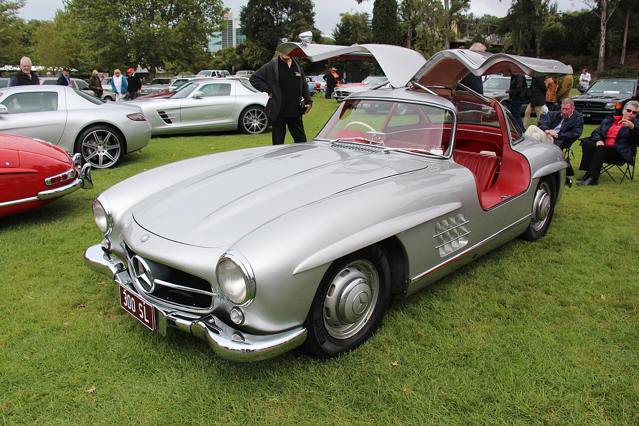 1956 Mercedes-Benz 300 SL Gullwing in silver with both doors raised and a red interior, front left three-quarter view on grass.