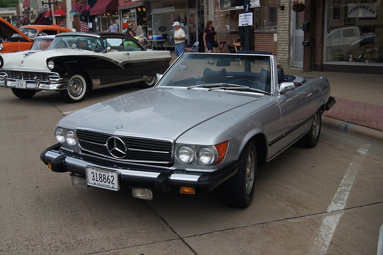 Silver 1975 Mercedes-Benz R107 SL convertible with its top down, front left three-quarter view on a town street at a car show.