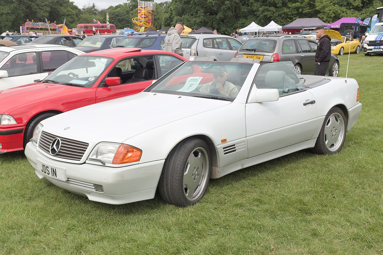 White Mercedes-Benz R129 SL with its roof down, front left three-quarter view on grass at an outdoor car show.
