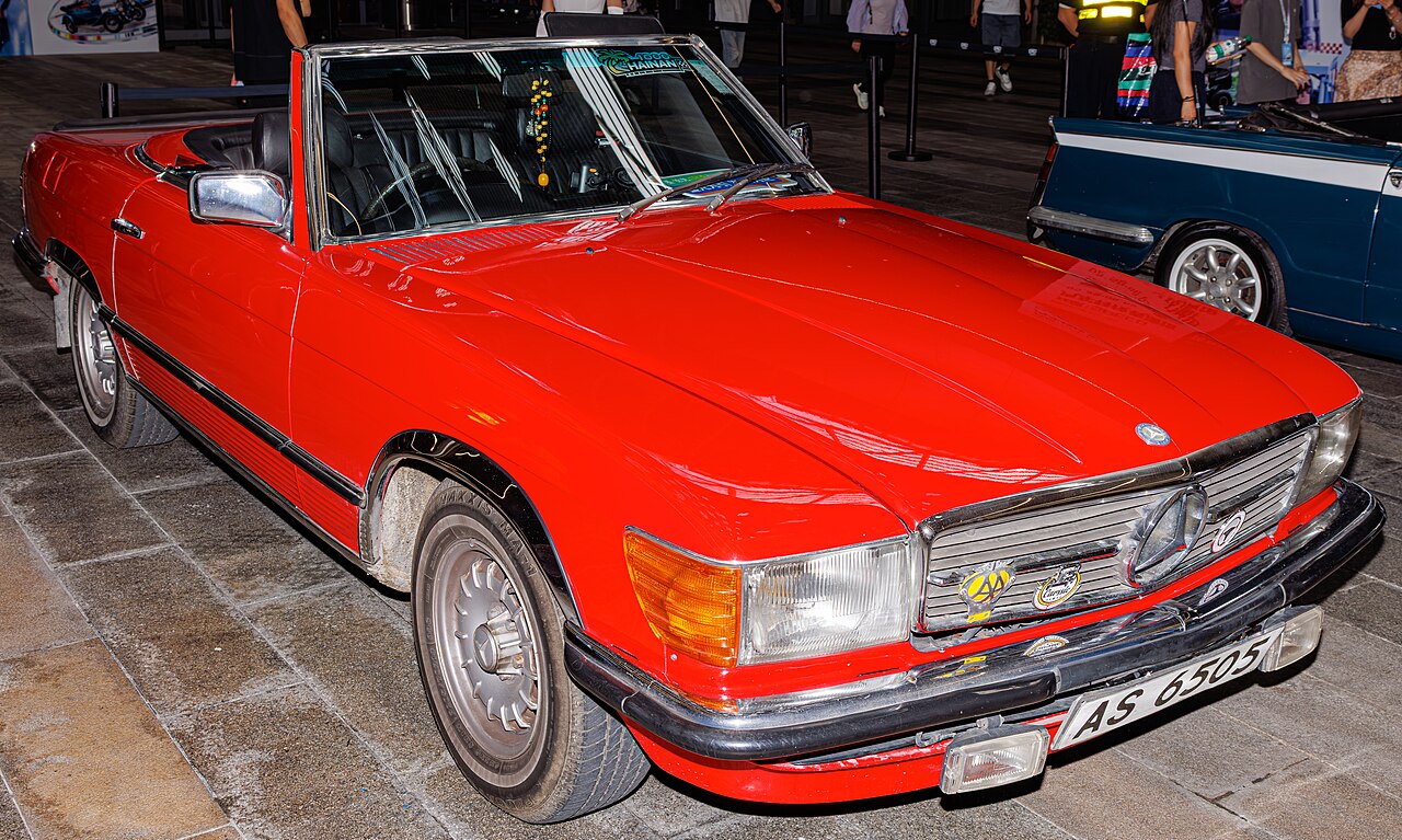Red Mercedes-Benz R107 SL roadster with its roof down, front three-quarter view parked on paving stones in Hong Kong.