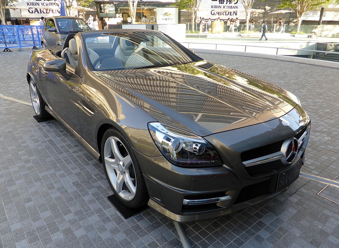 Grey-brown Mercedes-Benz SLK roadster with its roof down, front right three-quarter view on a paved plaza.