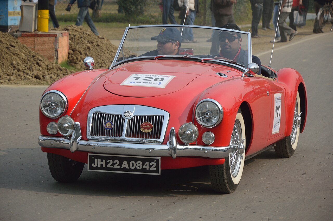 Red MGA 1600 Mk II roadster with wire wheels and whitewall tyres, two people aboard, front left view on a road during a rally.