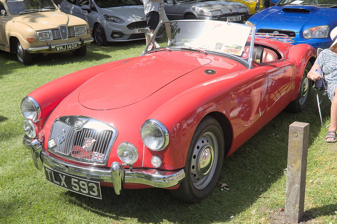 Red MGA roadster with its roof down, front left three-quarter view on grass at a classic car show.