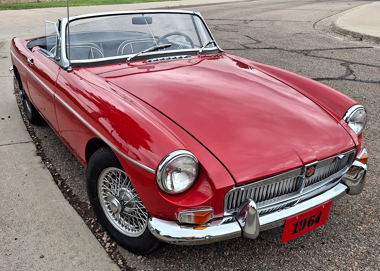 Red 1964 MG B roadster with wire wheels and its roof down, front right three-quarter view from above, parked at a kerb.