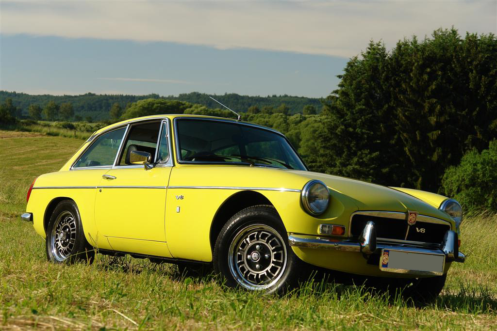 Yellow 1973 MG MGB GT V8 with chrome bumpers and V8 badges, front three-quarter view on a grassy field.