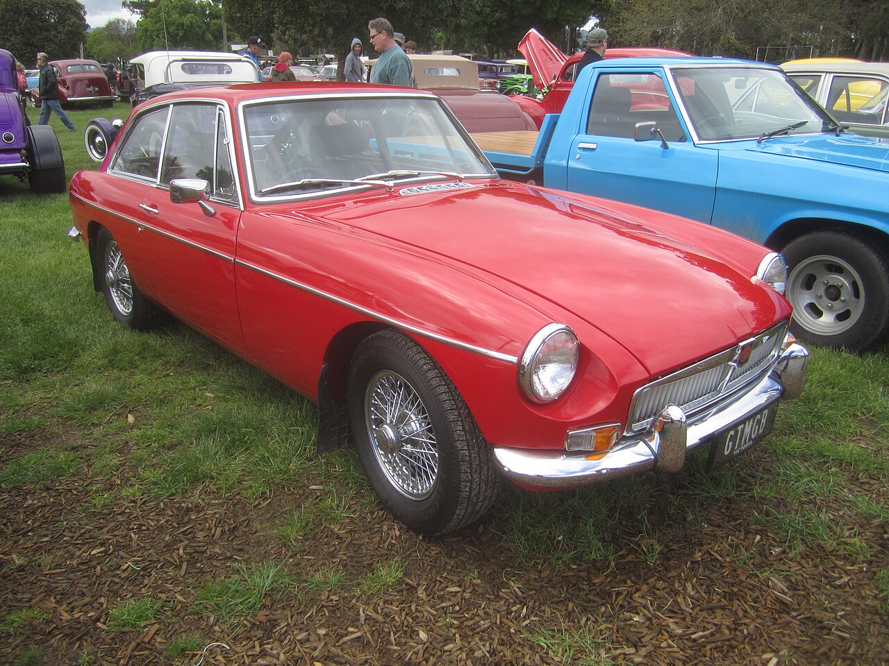 Red 1965 MG MGB GT with chrome bumpers and wire wheels, front three-quarter view on grass at a car show.