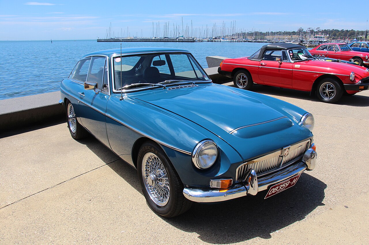 Blue 1968 MG MGC GT coupe with wire wheels, front three-quarter view on a waterfront promenade with boats behind.