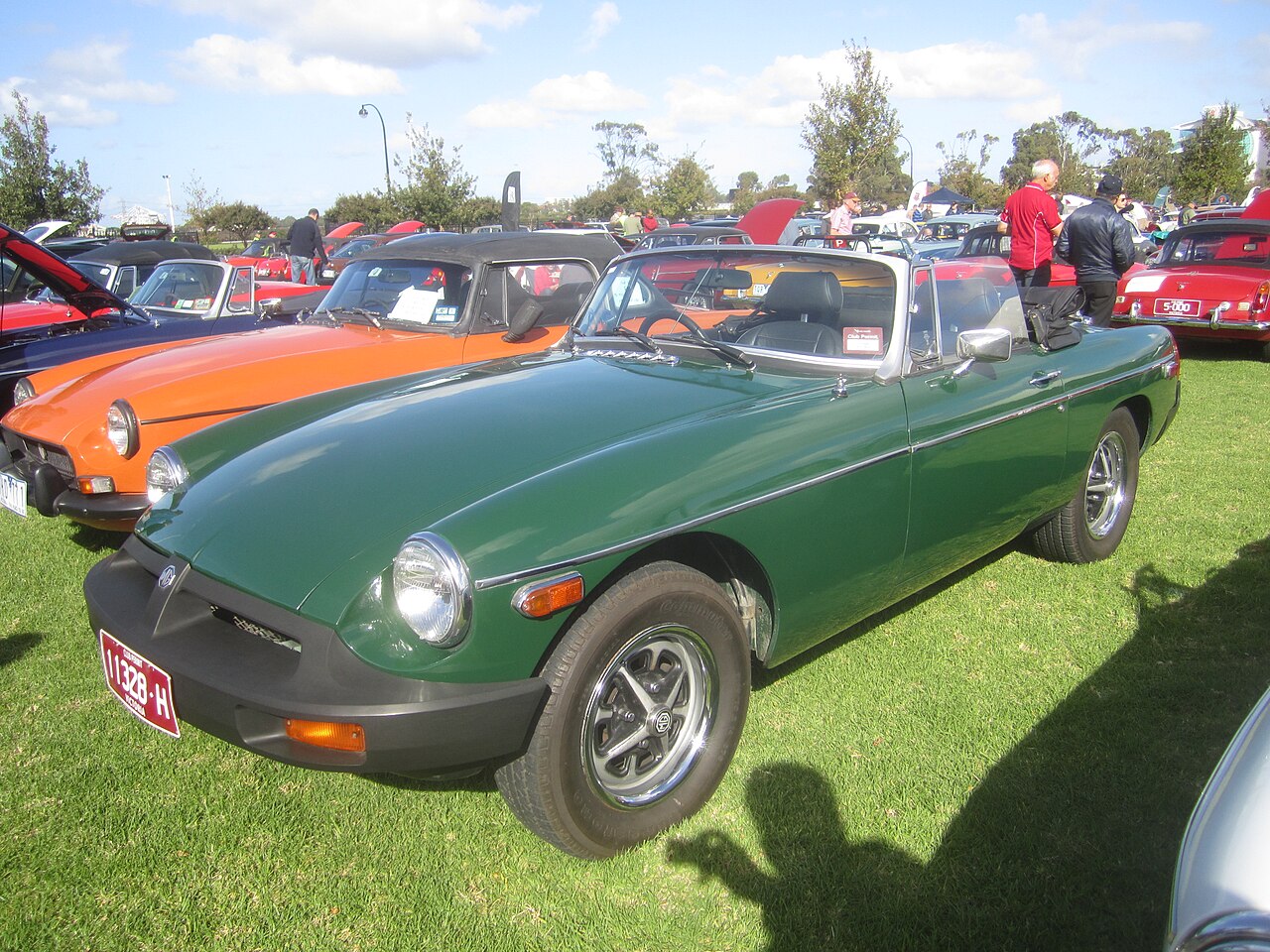 Green 1976 MG MGB roadster with black rubber bumpers and its roof down, front three-quarter view on grass at a car show.